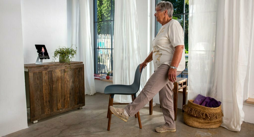 En eldre dame står på gulvet, med en hånd på stolen sin for å holde balansen, mens hun beveger på armer og ben. Foran seg på en skjenk har hun Berntsen Beinlaus, en iPad som hjelper henne med treningen.
