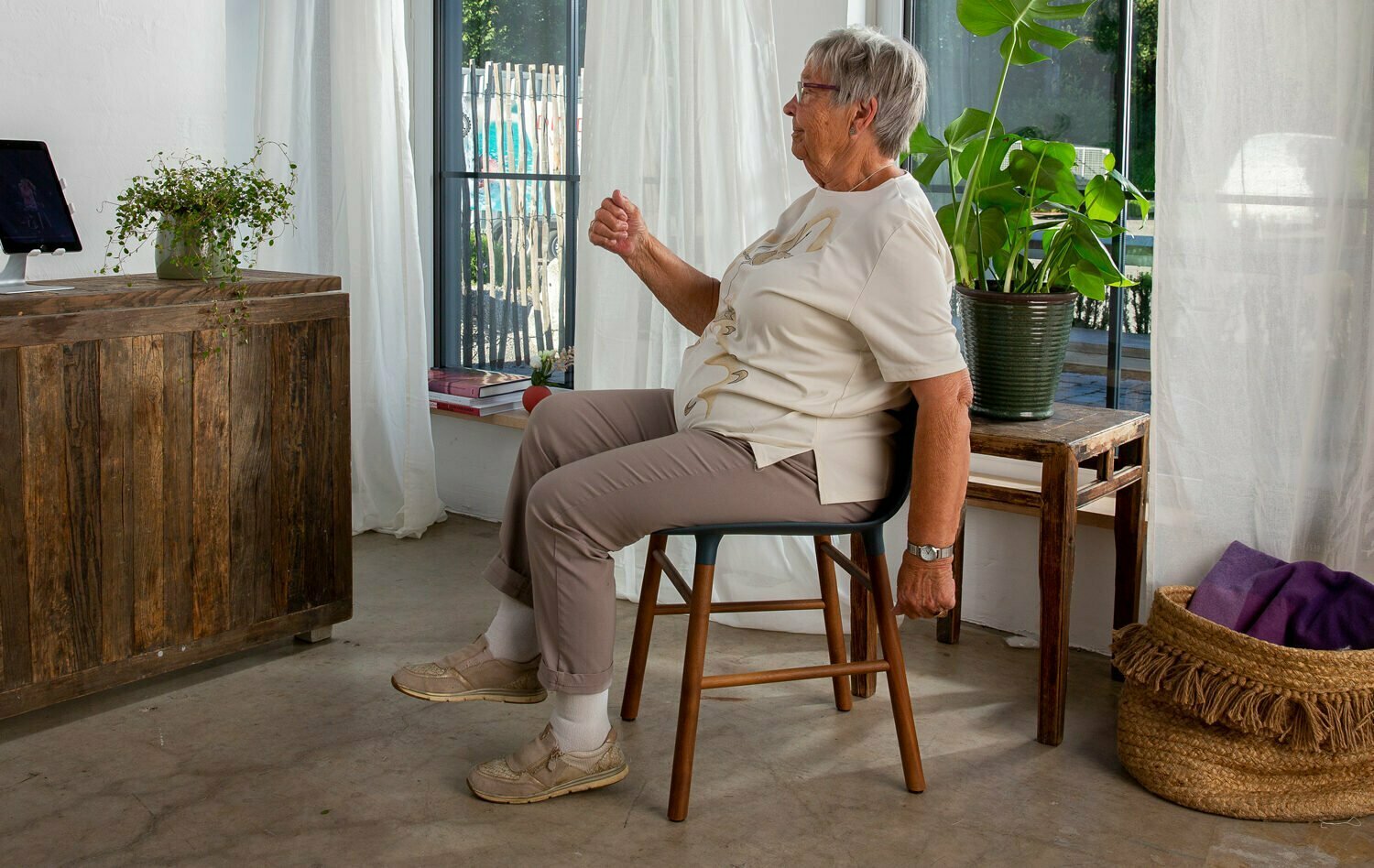 En eldre dame sitter på en stol i stua mens hun beveger på armer og ben. Foran seg på en sjenk har hun Berntsen Beinlaus, en ipad som hjelper henne med treningen.