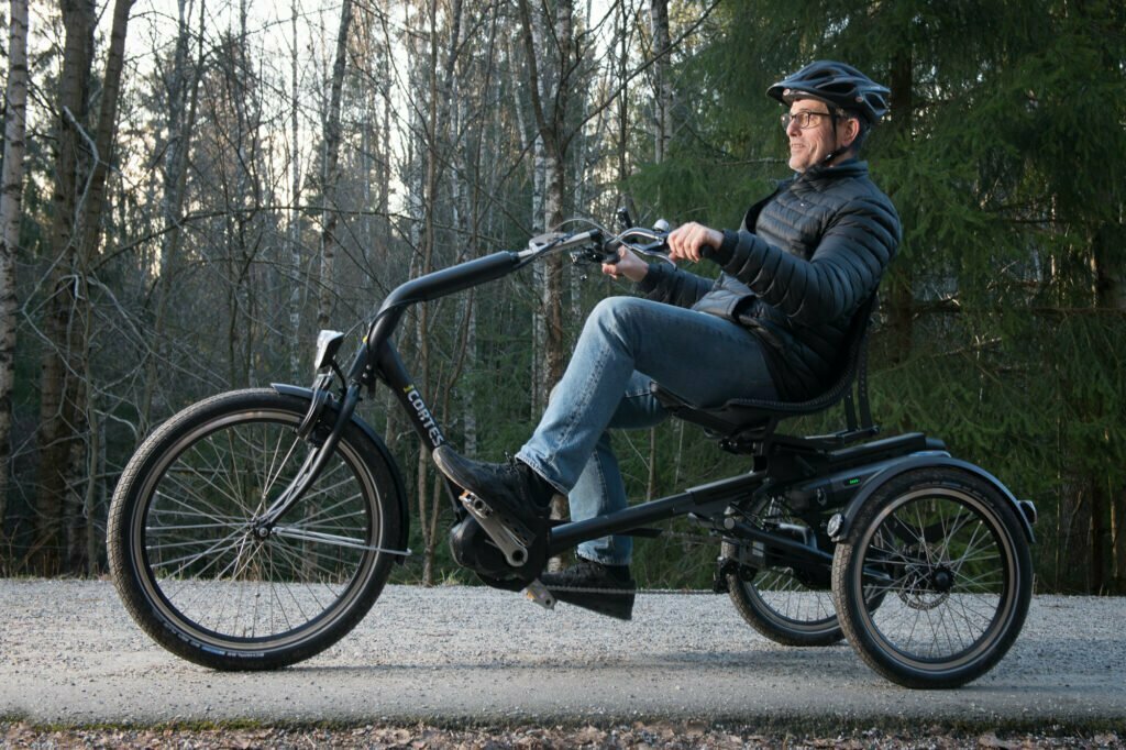 En mann i 60-årene sykler på trehjulssykkelen Cortes. Han sitter godt til rette i setet, mens han ser rett fremover. Han har på seg hjelm, tynn boblejakke og olabukse. Det er flere trær langs veien han sykler på.